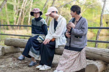 Üç kadın parkta bir kütüğe oturmuş, içlerinden biri bebek tutuyor. Manzara huzurlu ve rahatlatıcı, çünkü kadınlar birbirlerinin arkadaşlığından ve doğal çevreden hoşlanıyorlar.