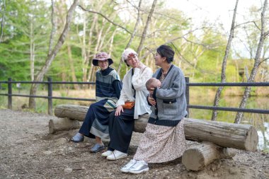 Parkta kütüğün üzerinde oturan üç kadın. İçlerinden biri bir bebek tutuyor. Manzara huzurlu ve rahatlatıcı.