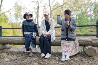 Üç kadın parkta bir kütüğün üzerinde oturuyorlar. İçlerinden biri bir bebek tutuyor. Manzara huzurlu ve rahatlatıcı.