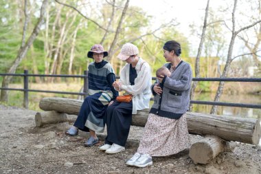 Üç kadın parkta bir kütüğe oturmuş, içlerinden biri bebek tutuyor. Manzara huzurlu ve rahatlatıcı, çünkü kadınlar birbirlerinin arkadaşlığından ve doğal çevreden hoşlanıyorlar.