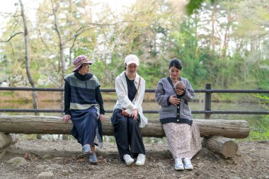 Parkta kütüğün üzerinde oturan üç kadın. İçlerinden biri bir bebek tutuyor. Manzara huzurlu ve rahatlatıcı.