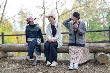 Parkta kütüğün üzerinde oturan üç kadın. İçlerinden biri bir bebek tutuyor. Manzara huzurlu ve rahatlatıcı.