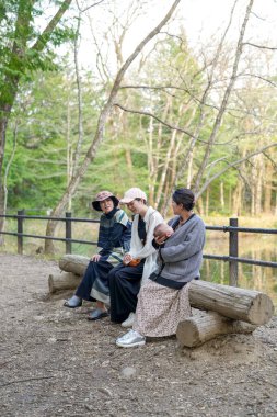Parkta kütüğün üzerinde oturan üç kadın. İçlerinden biri bir bebek tutuyor. Manzara huzurlu ve rahatlatıcı.