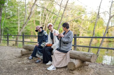 Üç kadın parkta bir kütüğe oturmuş, içlerinden biri bebek tutuyor. Manzara huzurlu ve rahatlatıcı, çünkü kadınlar doğal ortamda birbirlerine eşlik etmekten hoşlanıyorlar.