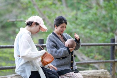 İki kadın yakınlarda otururken bir kadın bebeği besliyor. Sahne huzurlu ve huzurlu, kadınların bebekle bir anlık bir bağ kurmasıyla