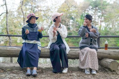 Üç kadın kütüğün üzerinde oturuyor, biri bebek tutuyor. Manzara huzurlu ve rahatlatıcı.