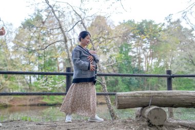 Bir kadın, siyah beyaz ceket giyerken taşıyıcı bir bebekle yürüyor. Sahne arka planda ağaçlar ve gölet olan bir parkta geçiyor. Manzara huzurlu ve sakin, tıpkı bir kadın gibi.