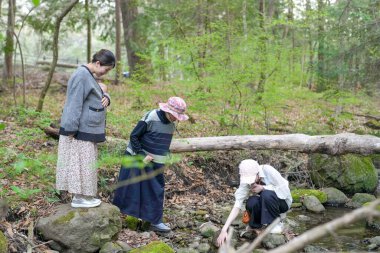 Üç kadın ve bir çocuk deredeki kütüğe bakıyorlar. Soldaki kadın süveter ve etek giyiyor.