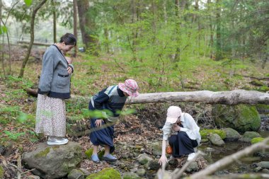 Üç kadın deredeki kütüğe bakıyor. Bir tanesi pembe şapka takıyor.