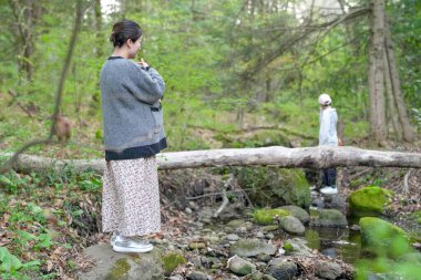 Bir kadın ormanda bir kütüğün üzerinde duruyor. Arkasından bir adam yürüyor. Kadın siyah beyaz bir ceket giyiyor.