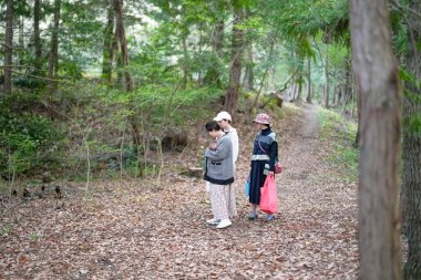 Üç kişi ormanda yürüyor, içlerinden biri bebek tutuyor. Sahne huzurlu ve huzurlu, grup doğa ile çevrili ve sessiz bir yürüyüşün tadını çıkarıyor.