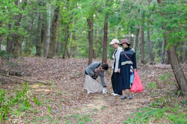 Üç kadın ormanda yürüyordu, biri bir şey almak için eğilmişti. Manzara huzurlu ve huzurlu, kadınlar doğa yürüyüşünden zevk alıyor.