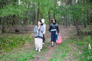 Üç kadın bir ormanda yürüyor, biri maske takıyor. Çantalar ve sırt çantaları taşıyorlar. Görüntü huzurlu ve huzurlu.