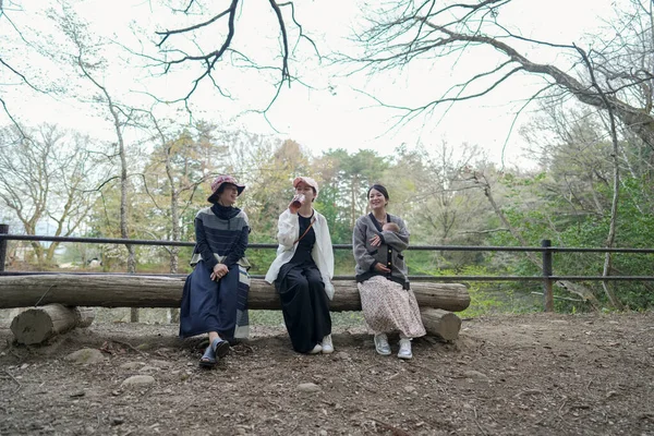 Parkta kütüğün üzerinde oturan üç kadın. Bir tanesi bardaktan içiyor. Manzara sakin ve huzurlu.