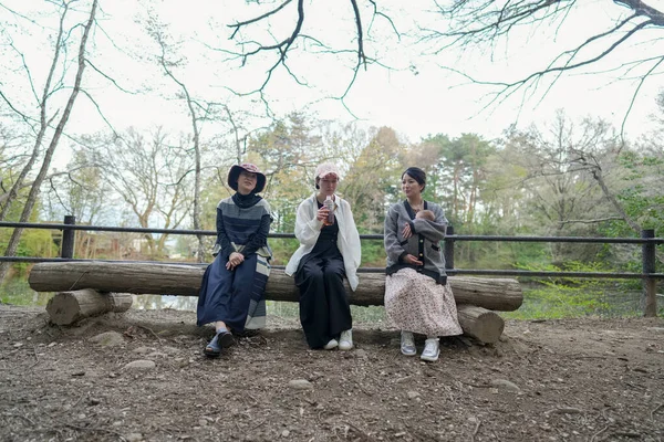 Parkta kütüğün üzerinde oturan üç kadın. İçlerinden biri şapka takıyor. Manzara huzurlu ve rahatlatıcı.