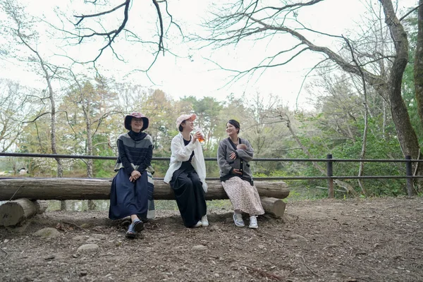 Parkta kütüğün üzerinde oturan üç kadın. İçlerinden biri şapka takıyor. Gülümsüyorlar ve birbirlerinin arkadaşlığından zevk alıyorlar.