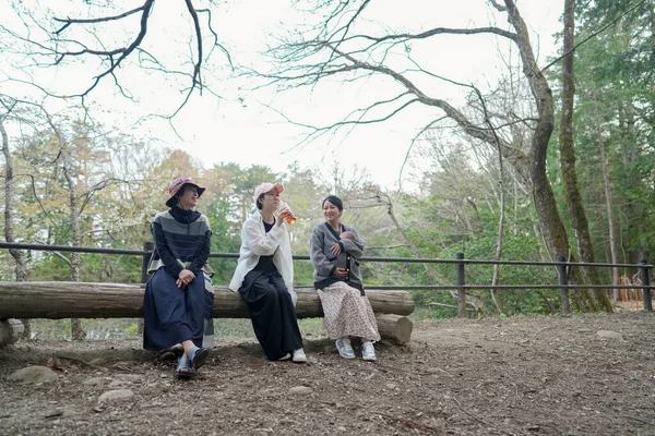 Parkta kütüğün üzerinde oturan üç kadın. İçlerinden biri bir içki tutuyor. Manzara sakin ve huzurlu.