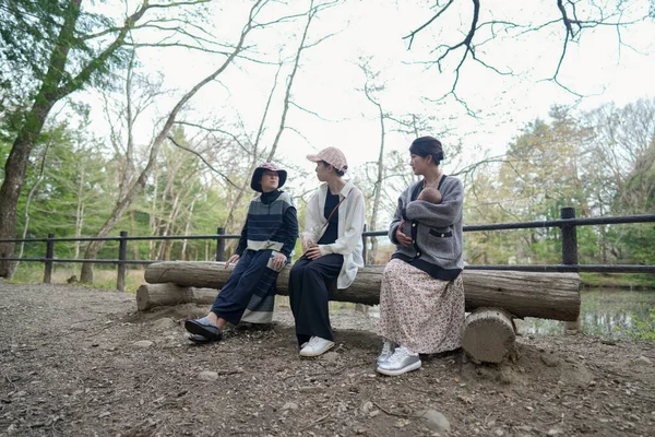 Parktaki bir kütüğün üzerinde üç kişi oturuyor. İçlerinden biri bir bebek tutuyor. Manzara huzurlu ve rahatlatıcı.