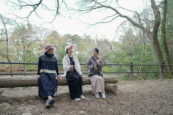 Parktaki bir kütükte üç kadın oturuyor. İçlerinden biri bir bebek tutuyor. Manzara huzurlu ve rahatlatıcı.