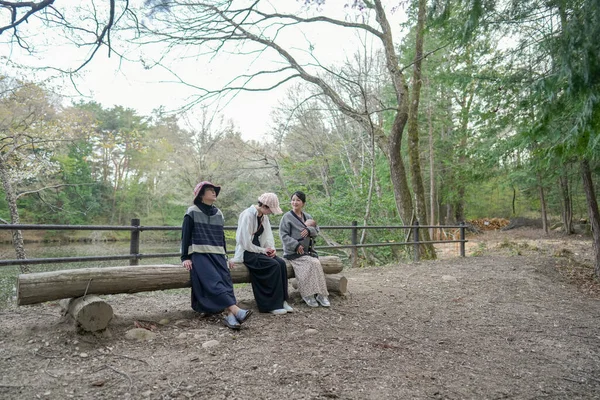 Parktaki bir kütükte üç kadın oturuyor. İçlerinden biri şapka takıyor. Manzara huzurlu ve rahatlatıcı.