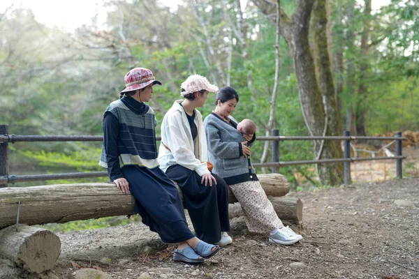 Bir grup kadın ormandaki bir kütüğe oturmuş, içlerinden biri bebek tutuyor. Manzara huzurlu ve huzurlu, çünkü kadınlar birbirlerine eşlik etmekten ve doğal çevreden hoşlanıyorlar.