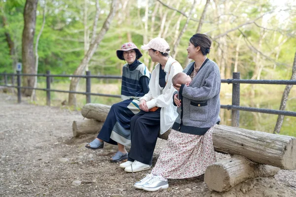 Üç kadın ormanda bir kütüğün üzerinde oturuyor, biri bebeği tutuyor. Manzara huzurlu ve huzurlu, çünkü kadınlar doğal ortamda birbirlerine eşlik etmekten hoşlanıyorlar.