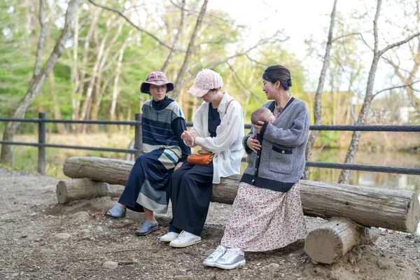 Üç kadın parkta bir kütüğe oturmuş, içlerinden biri bebek tutuyor. Manzara huzurlu ve rahatlatıcı, çünkü kadınlar birbirlerinin arkadaşlığından ve doğal çevreden hoşlanıyorlar.