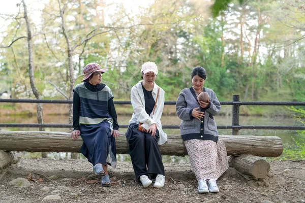 Parkta kütüğün üzerinde oturan üç kadın. İçlerinden biri bir bebek tutuyor. Manzara huzurlu ve rahatlatıcı.