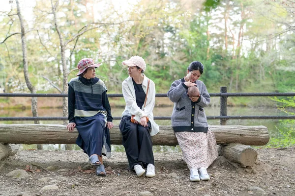 Parkta kütüğün üzerinde oturan üç kadın. İçlerinden biri bir bebek tutuyor. Manzara huzurlu ve rahatlatıcı.