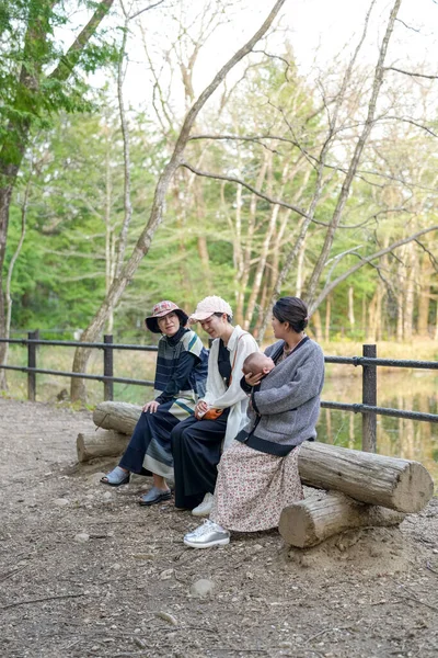 Parkta kütüğün üzerinde oturan üç kadın. İçlerinden biri bir bebek tutuyor. Manzara huzurlu ve rahatlatıcı.