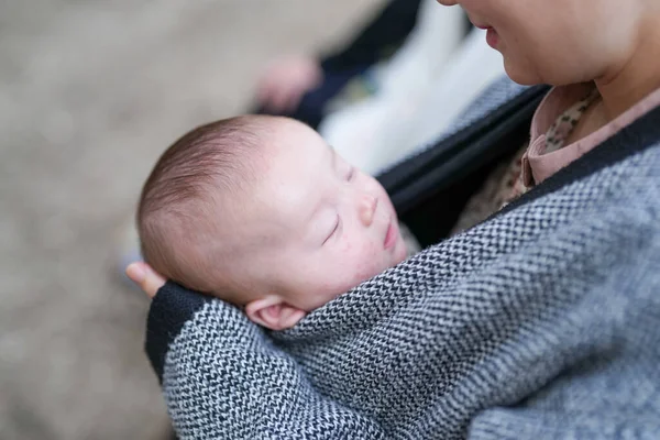 Bir kadın uyuyan bir bebeği kollarında tutuyor. Bebek bir battaniyeye sarılmış ve çok yorgun. Kadın gri ve siyah bir ceket giyiyor ve yerde oturuyor.