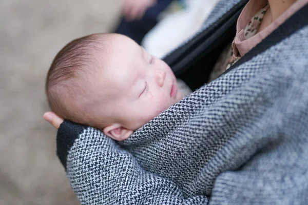 Bir kadın uyuyan bir bebeği kollarında tutuyor. Bebek bir battaniyeye sarılı ve bir şapka giyiyor. Kadın gri bir ceket ve siyah bir gömlek giyiyor. Sıcaklık ve rahatlık kavramı