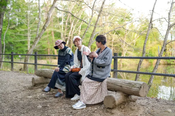 Üç kadın parkta bir kütüğe oturmuş, içlerinden biri bebek tutuyor. Manzara huzurlu ve rahatlatıcı, çünkü kadınlar doğal ortamda birbirlerine eşlik etmekten hoşlanıyorlar.