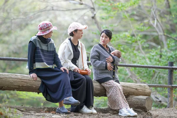 Üç kadın ormanda bir kütüğün üzerinde oturuyor, biri bebeği tutuyor. Manzara huzurlu ve huzurlu, çünkü kadınlar doğal ortamda birbirlerine eşlik etmekten hoşlanıyorlar.