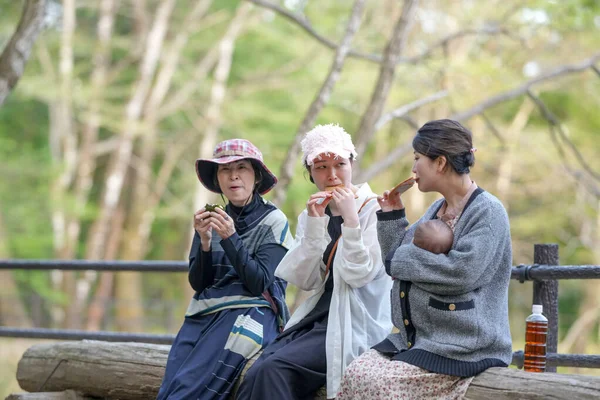 Üç kadın bir bankta oturuyor, biri bebeği tutuyor. Manzara huzurlu ve rahatlatıcı.