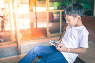 Genç Asyalı çocuk cep telefonları oynarken ahşap bir sandalyede oturan 