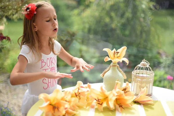 A little girl at the table on terrace. Beautiful yellow lilies are on the table. Girl wears pink flower in the head. She is very emotional and smily. There\'s a yellow striped tablecloth.