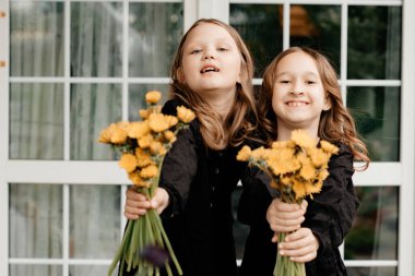 Siyah elbiseli iki kız ellerinde çiçeklerle birbirlerine gülümsüyorlar. Lüks, parlak saçlar. Yazın iyi eğlenceler ve rahatla. Çocuk stili ve moda kavramı. Uzayı kopyala Tonlama.