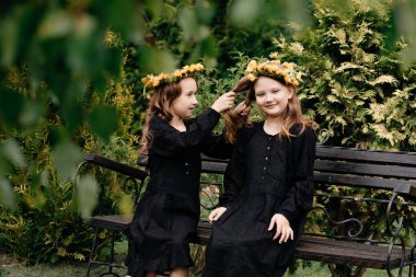 Siyah elbiseli küçük kızlar bankta oturuyorlar. Temiz havada rahatla ve eğlen. Çocuk stili ve moda kavramı. Arka planda çiçekler.  
