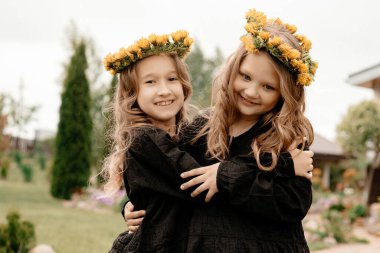 Kızlar başlarının üzerinde karahindiba çelenkleri ile gülümseyerek birbirlerine sarılırlar. Arka planda çimenler var. Spor ve pozitif kavramı. Uzayı kopyala Tonlama.