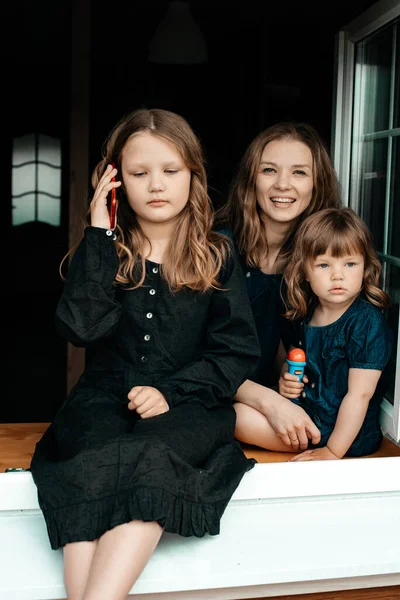 年轻迷人的妈妈和两个穿着黑色衣服的小女孩坐在窗台上 一个小女孩在讲电话 暑假无忧无虑的童年 — 图库照片