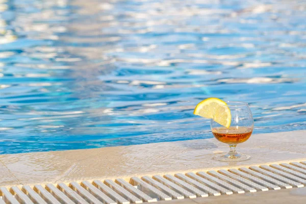 Glass of cognac, a slice of lemon, blue pool water as an attribute of a luxurious holiday