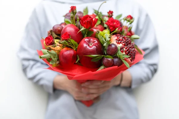 Edible bouquet consisting of pomegranate, apples, plums and scarlet roses in hands of woman on white background