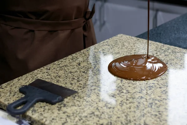 Chocolatier in a brown apron pours liquid chocolate on a granite table