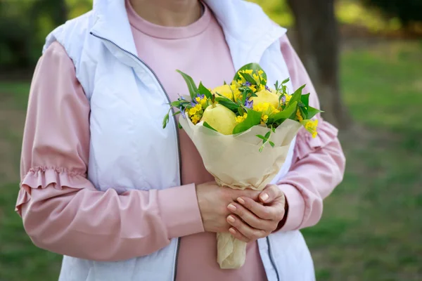 Cute bouquet of flowers and fruits in the hands of a young woman on a background of green grass