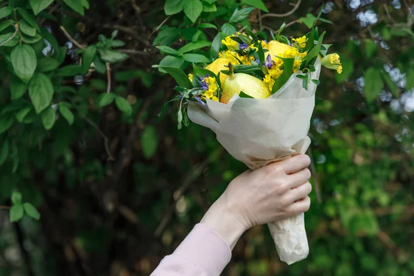 Bouquet of yellow and blue flowers and fruits in the hand of a young woman on a background of green foliage