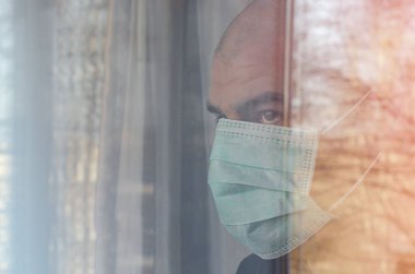 Beyaz adam koruyucu maskeli pencereden dışarı bakıyor. Karantina sırasında kendini izole etme.