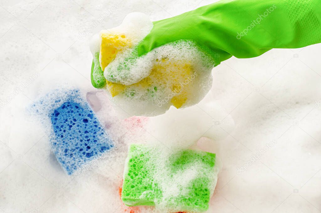 Hand in protective glove holding a soapy sponge on foam background. Housewifery concept.