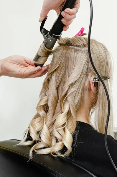 Stylist making a hairdo with hair curler for blond woman after hair dyeing. Beauty salon concept.