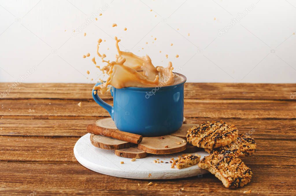 Blue mug with splashing coffee and cookies on wooden background.